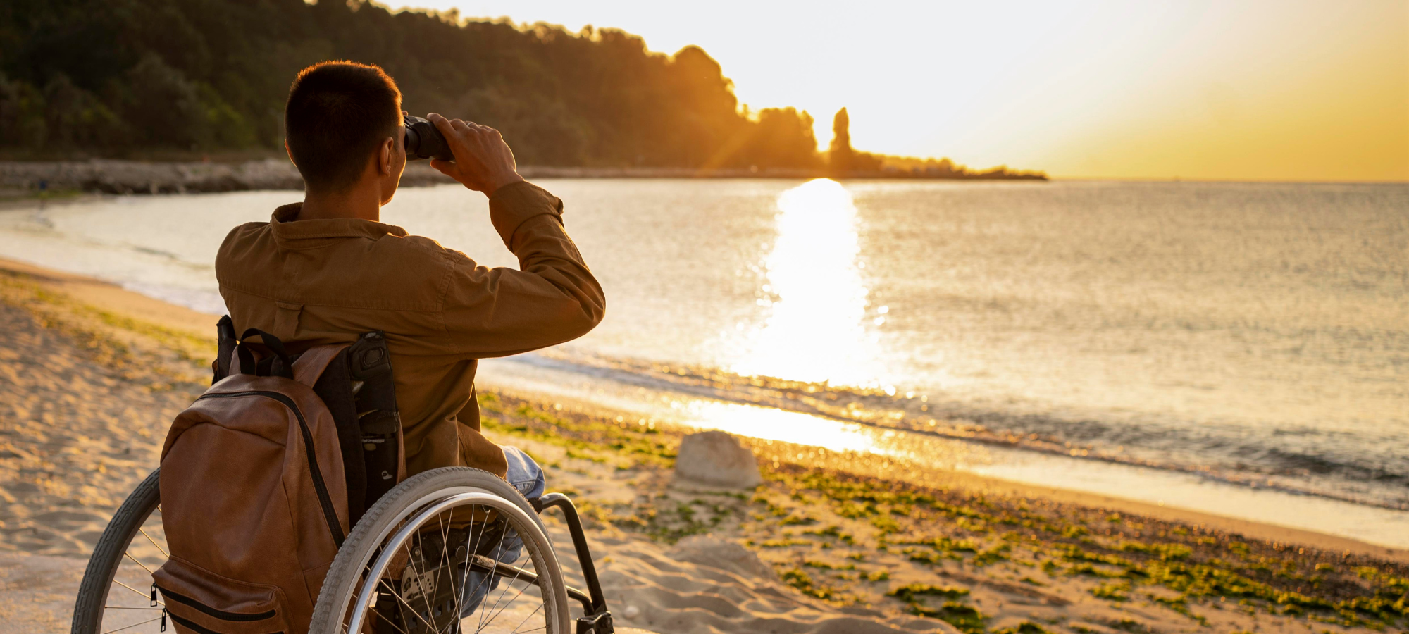 Homme en fauteuil roulant observant le coucher de soleil sur une plage, illustrant l'autonomie malgré le handicap